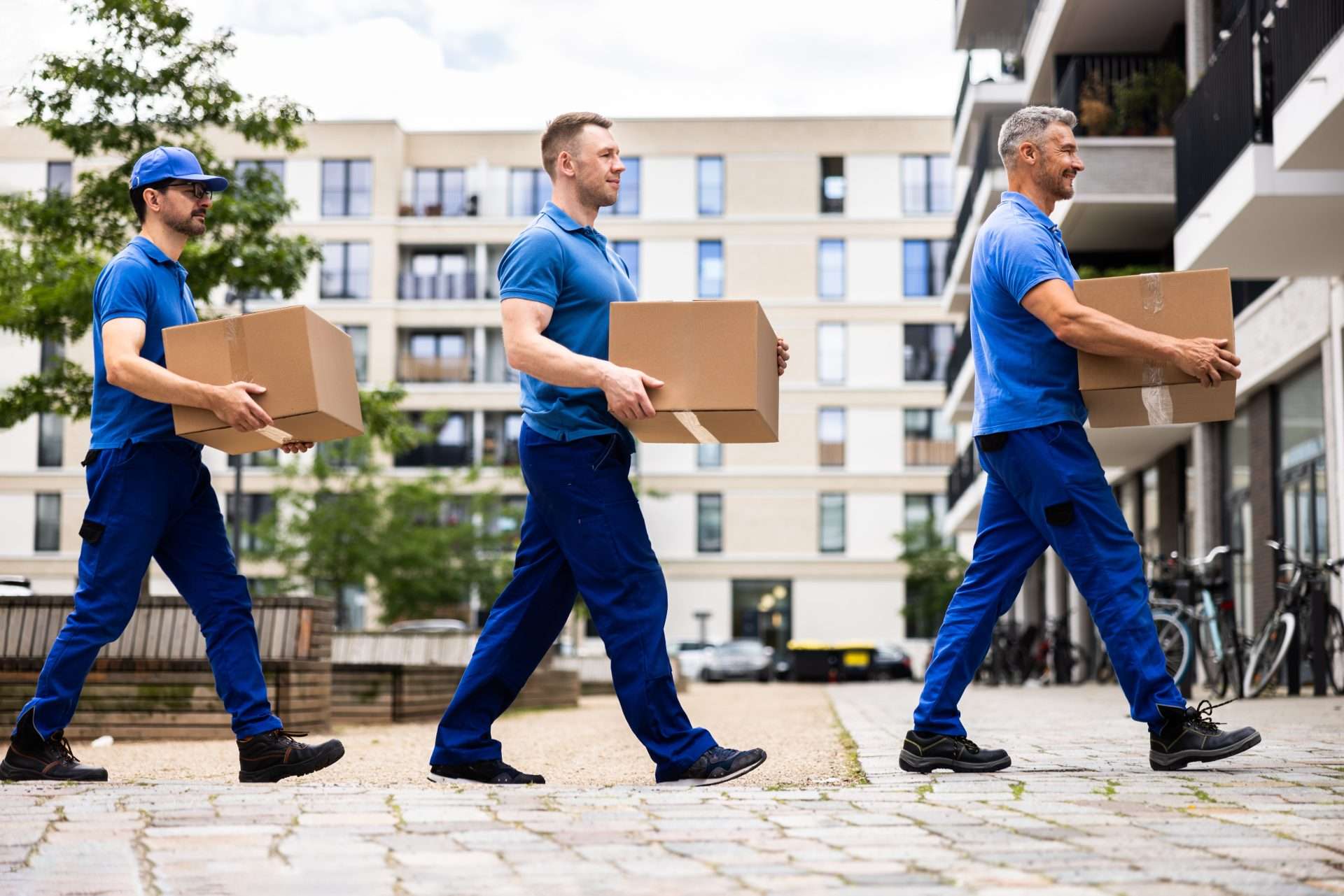 Three movers dressed in blue, walking in a straight line while carrying one box each.