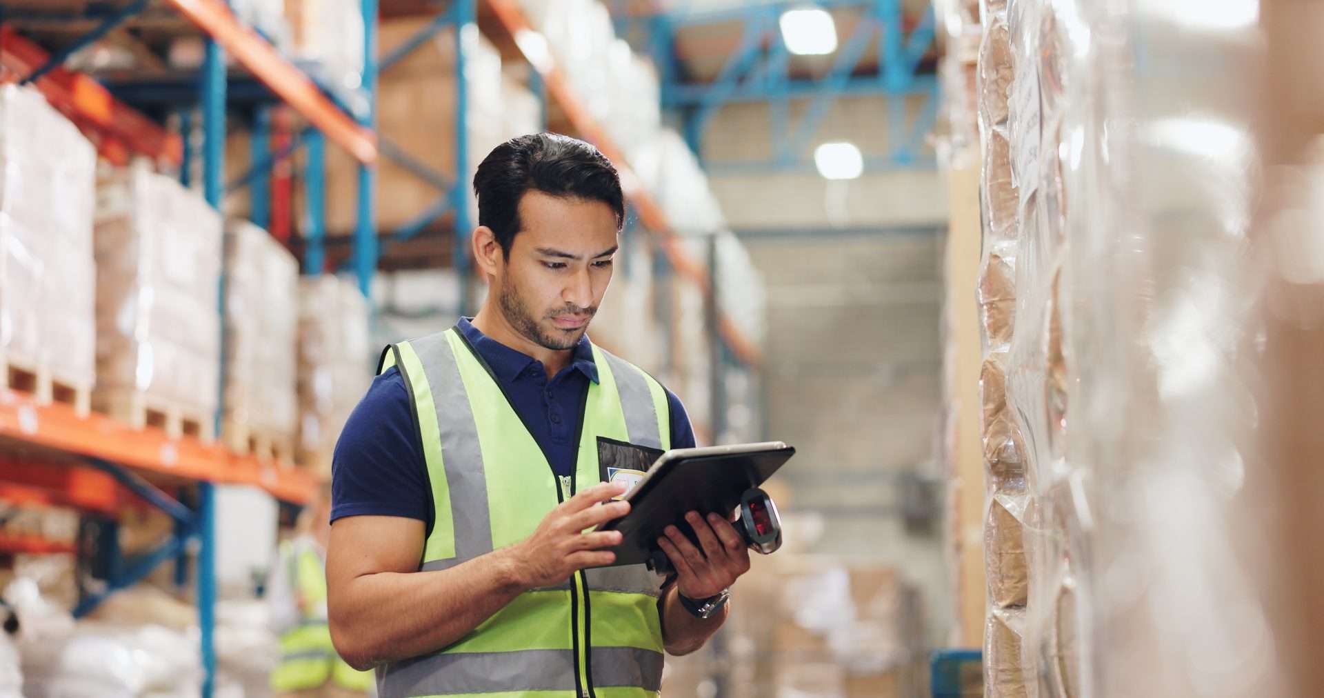 A warehouse worker in a yellow safety vest holding a scanner and a tablet