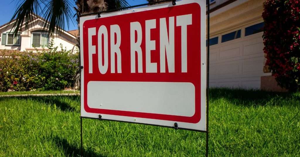 A red “For Rent” sign in the grass in front of a single-family home